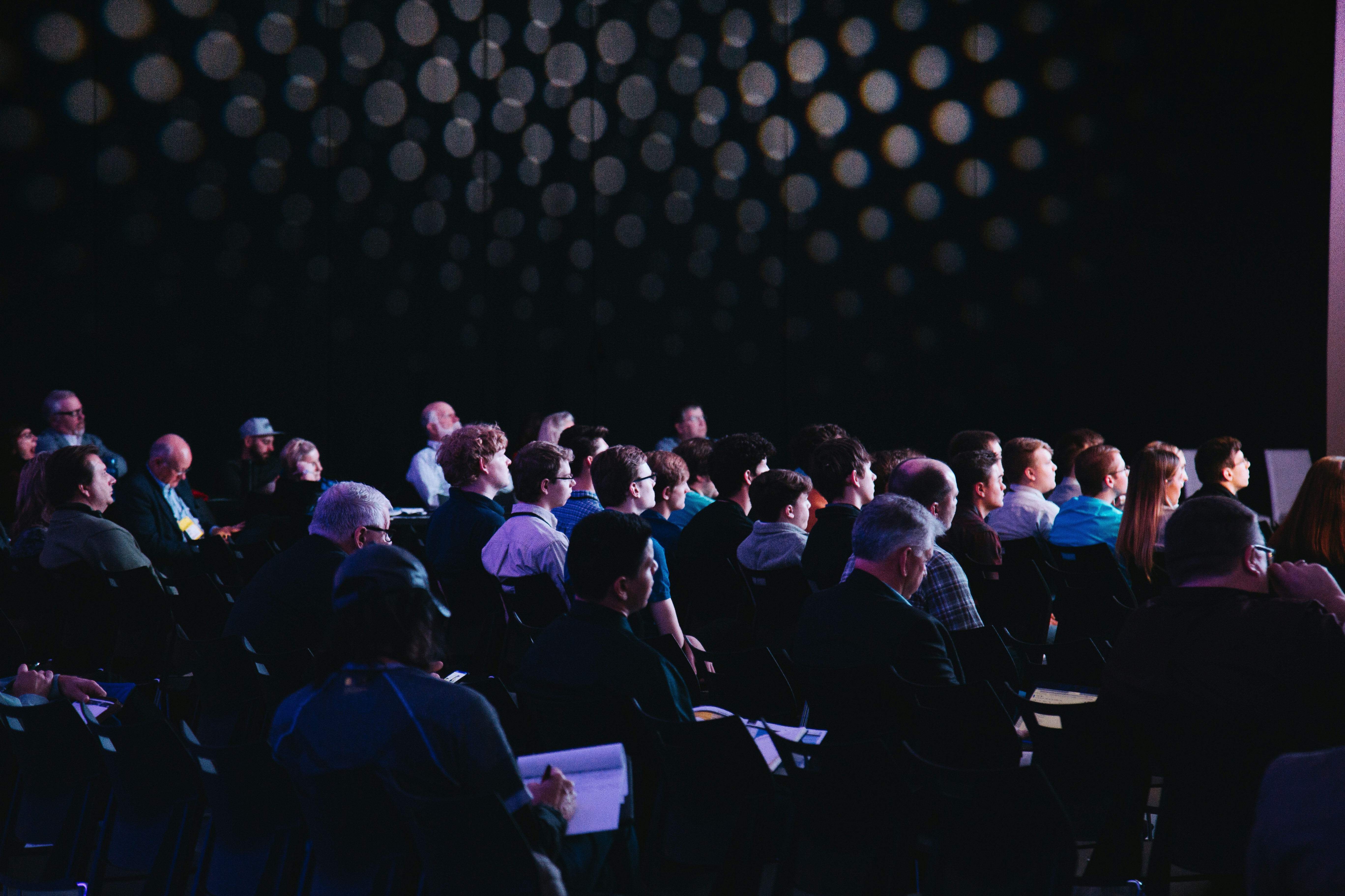 Engaged audience at a live event venue seated in rows under atmospheric purple stage lighting